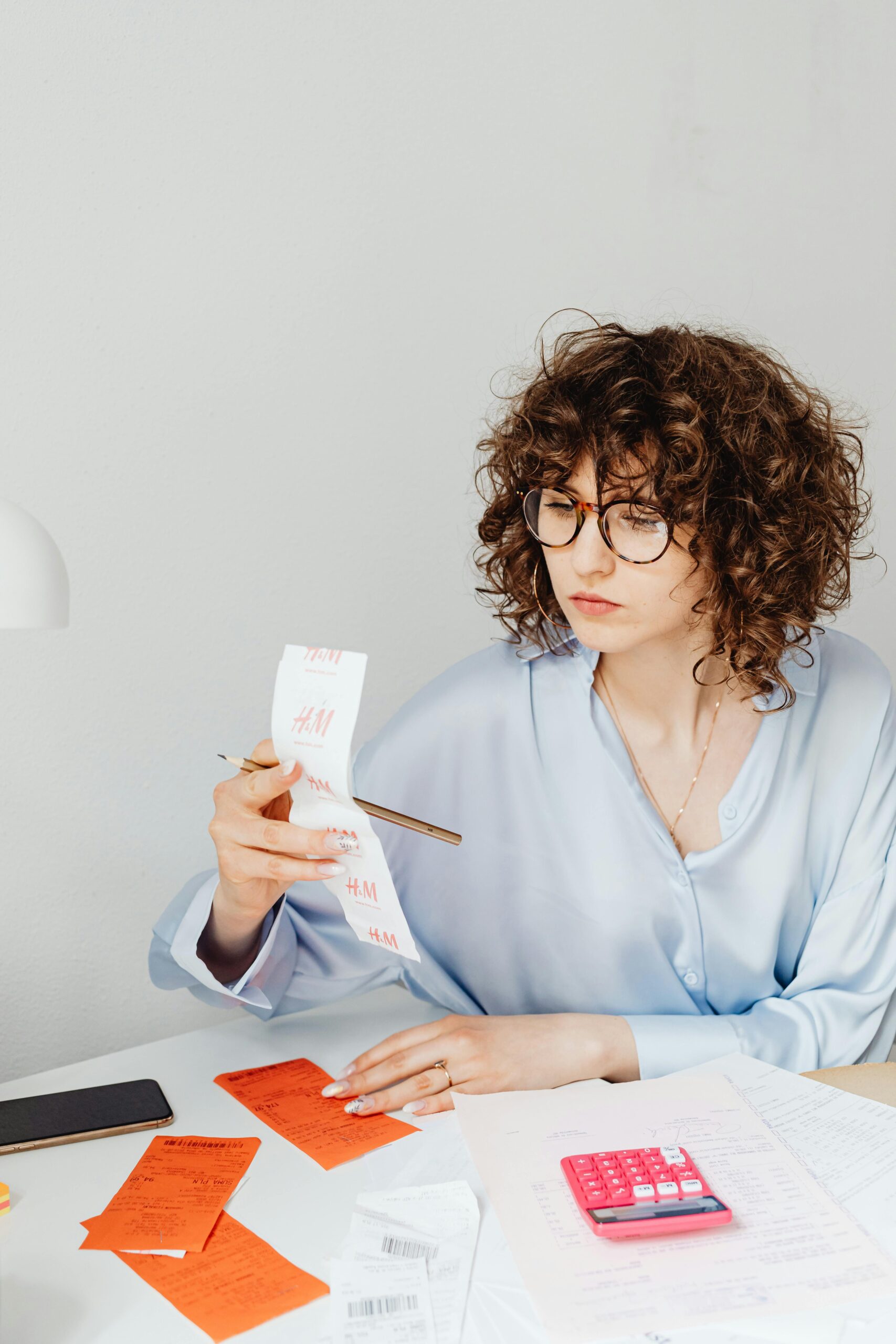 Curly-haired woman examining receipts with a calculator at her desk.
